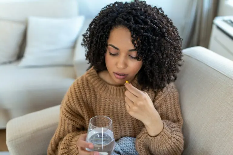 a young black woman sitting on the couch, taking a pill and a glass of water